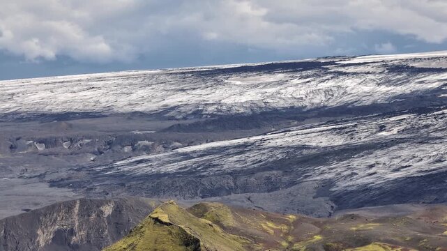 Experience a breathtaking aerial pan left across Iceland's rugged landscape. Witness green mossy hills and an vast, intimidating  glacier in the distant.