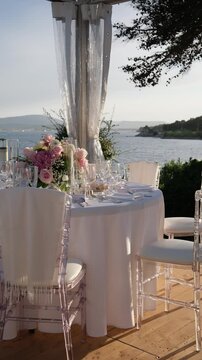 Beautifully decorated wedding table with pink flowers, crystal glasses, and transparent chairs standing on a wooden terrace overlooking the sea during a picturesque and romantic sunset. Vertical video