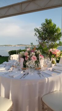 Beautifully decorated round table with a white tablecloth, pink rose bouquets, and elegant tableware set for a wedding reception or gala dinner by the sea during a sunny summer day. Vertical video