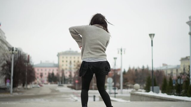 Young woman dances freely in urban setting her movements fluid and expressive. Woman wears light gray knit sweater over white shirt with black leggings and boots as snow covers ground