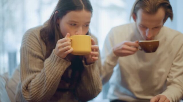 Close up of two people drinking beverages one holding yellow mug and other with brown cup in soft indoor lighting. Both people are engaged together at table setting with cups and saucers