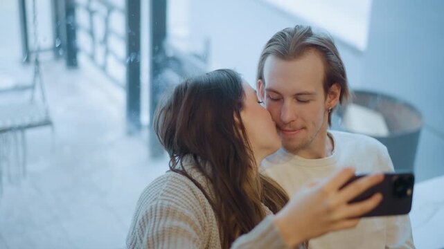 Close up of young couple taking selfie together indoors with smartphone. Woman holds phone while man leans in towards her both engaged and smiling. Soft natural light fills bright modern