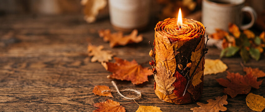 Handmade candle decorated with autumn leaves burning on wooden table. Cozy fall home decor with hot drink and dry foliage. Seasonal autumn atmosphere and hygge concept.