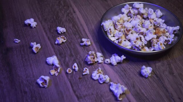 Close-up overhead view of a hand grabbing butter and chocolate-coated popcorn from a bowl, causing pieces to spill onto a wooden table during movie night.
