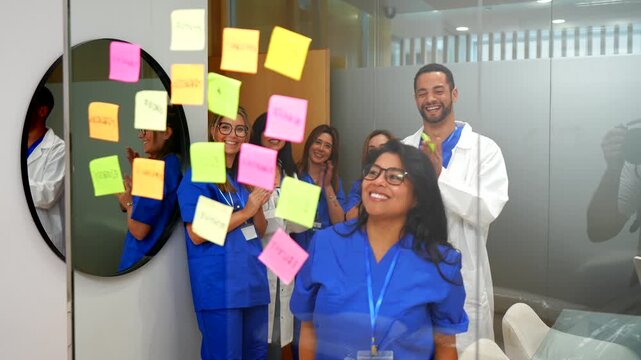 Diverse doctors and nurses celebrate a successful brainstorming session in a modern hospital office as a woman places the final colorful sticky note on a glass wall while colleagues applaud