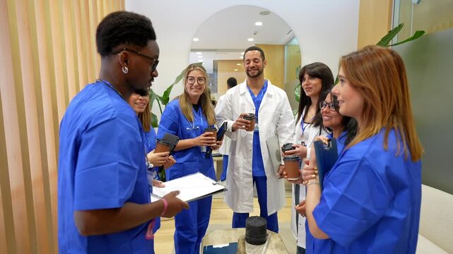 Multicultural group of doctors and nurses in scrubs and lab coats taking a break, drinking coffee, and talking together in the corridor of a modern medical center or private clinic