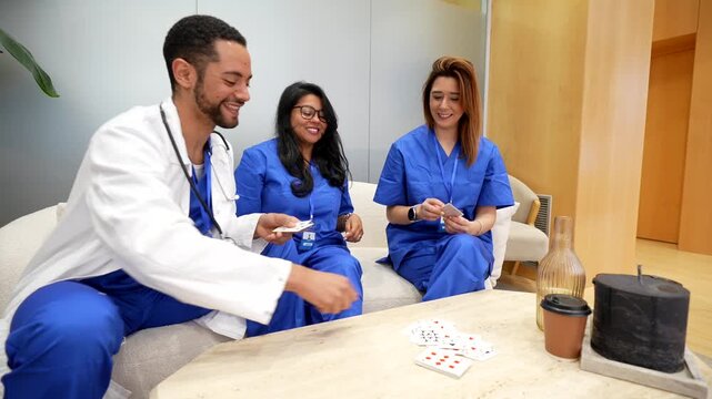 Diverse young doctors and nurses in a hospital staff room laughing and playing cards on break, sharing camaraderie, relaxation and friendly bonding between colleagues
