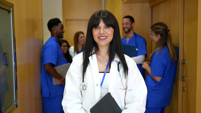 Portrait of a confident young female doctor in a white coat holding a tablet and smiling at the camera, while her diverse medical team of nurses talks in the background of a hospital corridor