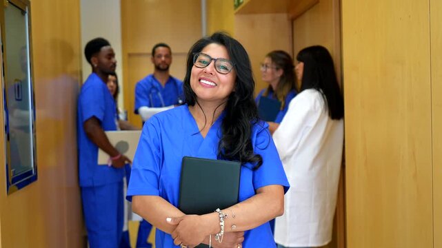 Portrait of a proud hispanic female nurse wearing blue scrubs and glasses, smiling at the camera while standing in a busy hospital hallway with her diverse medical team in the background