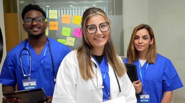 Professional and diverse medical team smiling at the camera in a modern hospital hallway. A female doctor in a white coat and two nurses in blue scrubs represent healthcare professionals at work