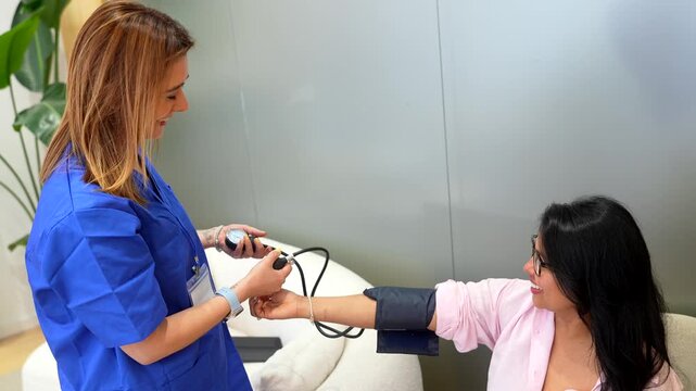 Female doctor in blue uniform using a tonometer to check the blood pressure of a smiling female patient during a routine medical examination in a modern hospital waiting room
