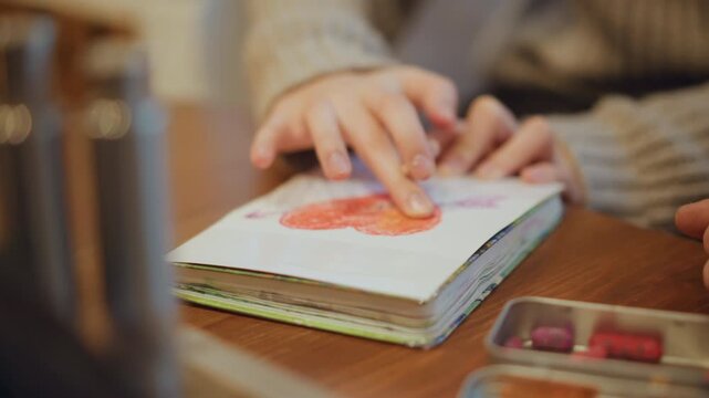 Close up of hands interacting with book and crayons on table. Hands press down red heart drawing in sketchbook while other hand rests nearby surrounded by art supplies including metal tin