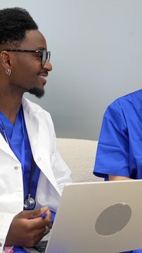 Two multiethnic medical professionals, an african american doctor and a caucasian nurse, sitting together in a clinic's staff room discussing patient records and diagnosis on a laptop computer