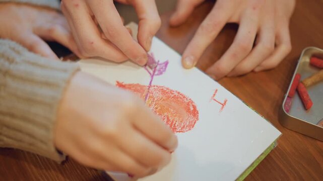 Close up of hands drawing with pastels on paper creating colorful artwork. Two people collaborate to draw heart and arrow design using red orange purple crayons in soft lighting. Hands work