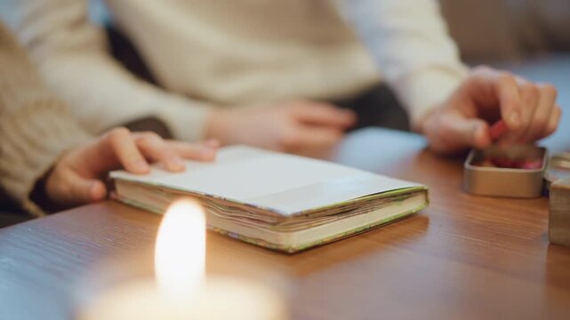 Close up of lit candle on wooden table with notebook and hands in background. Notebook pages are stacked showing colorful edges under soft lighting. Hands rest near book while another hand