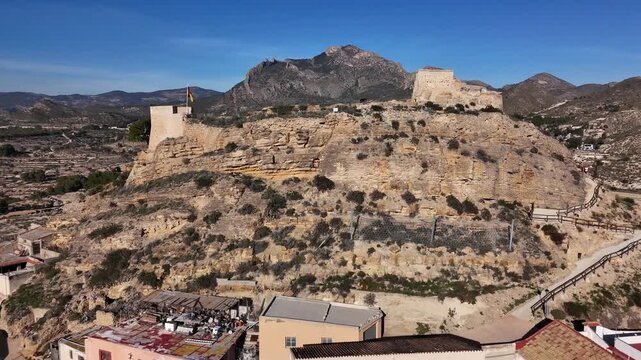 Drone view of the historic Busot Castle on a rocky hill with the Spanish flag, surrounded by rugged mountains and the scenic landscape of Alicante, Spain.