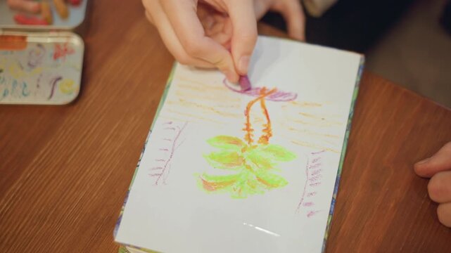 Close up of young girl coloring on white paper with pastels creating colorful abstract lines and shapes. Her hands hold crayon steadily as girl adds details to existing drawing focusing