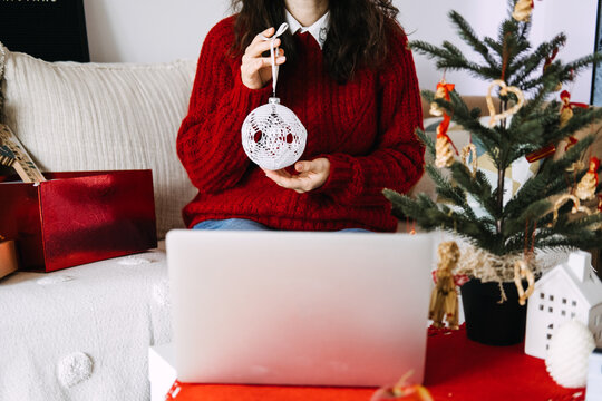 Woman shows lace ornament toward laptop screen while sitting near decorated Christmas tree. charity video call december, spreading goodwill online, connecting isolated people