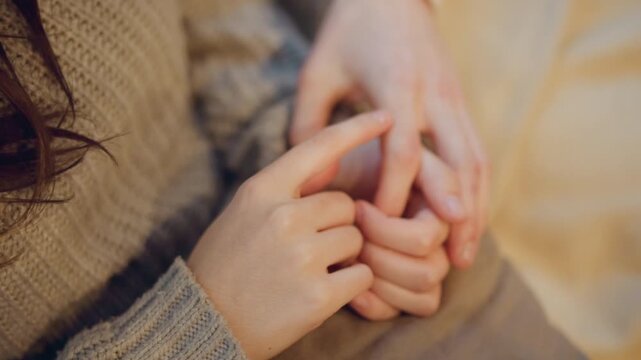 Close up of young woman gently holding another woman hand with soft lighting and warm tones. interaction shows tender connection between two hands one wearing textured knitwear. Background