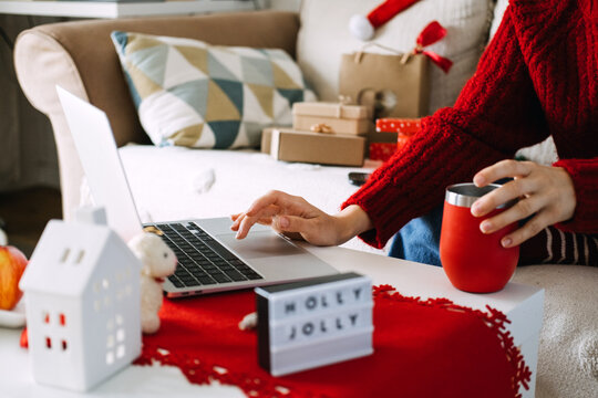 Woman uses laptop while holding mug beside wrapped parcels and HOLLY JOLLY sign at home. Handmade shop holiday orders, side hustle december income, solo entrepreneur workspace home