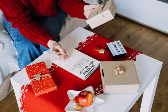 Woman holds wrapped box while writing in notebook with CHRISTMAS SHOPPING LIST beside HOLLY JOLLY sign. Christmas shopping list budget, holiday spending plan, planning christmas expenses