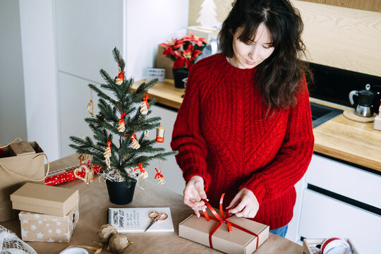 Woman ties ribbon on kraft box beside notebook with CHRISTMAS SHOPPING LIST in kitchen. Balancing work christmas season, holiday time management concept, juggling work and holidays