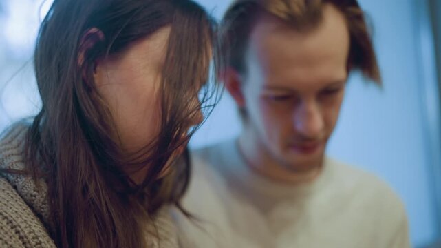 Close up of two people woman with long hair and man standing together in soft indoor lighting. subjects appear focused on something out of frame while interacting closely. Background