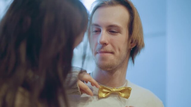 Close up of young woman carefully placing and adjusting golden bow tie on neck of man with light brown hair. interaction is gentle focusing on precise hand movements as woman positions