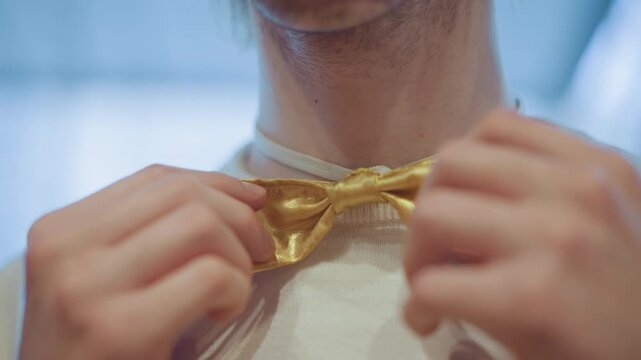 Close up of man adjusting his golden bow tie around neck while wearing white shirt with soft lighting and indoor background closeup bowtie neckwear fabric
