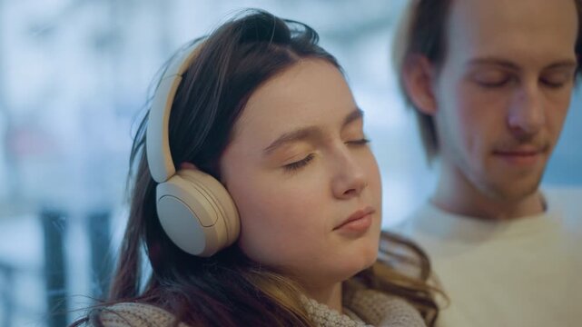 Close up of woman wearing beige over ear headphones with eyes closed man in white shirt visible behind her. Soft lighting highlights natural textures and calm expressions throughout scene