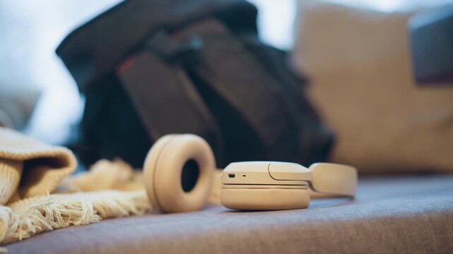 Close up of white wireless headphones resting on soft surface with natural light. black backpack and beige blanket are visible in softly blurred background suggesting travel or daily life