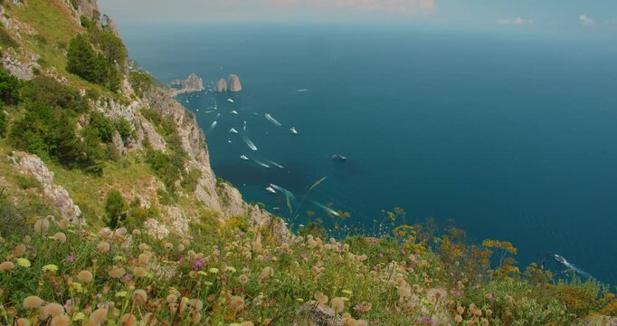 Steep coastal cliffs overlooking the sea and Faraglioni sea stacks filled with boats in Capri Italy