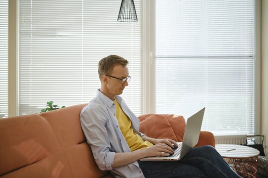 A retired man sits comfortably on a couch and writes his memoirs on laptop in a well-lit living room. Sunlight filters through the blinds, creating a warm and creative atmosphere