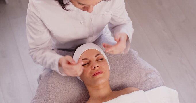 Woman enjoying face lifting massage, lying on couch, wrapped in towel, having pampering procedures in spa salon, keeping eyes closed. Professional cosmetologist doing facebuilding massage