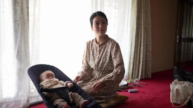 A woman turns toward an infant and gestures gently after her hairstyle has been arranged Natural window light fills the traditional interior