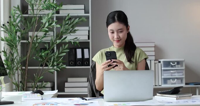 Young asian woman sitting at her desk, using a smartphone while a laptop and monitor are visible, reflecting concepts of remote work, technology, and productivity in a contemporary office setting