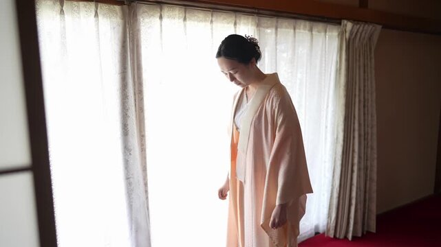 A woman stands in a pale peach kimono underlayer near a bright window Her finished updo and relaxed posture suggest ongoing formal dressing