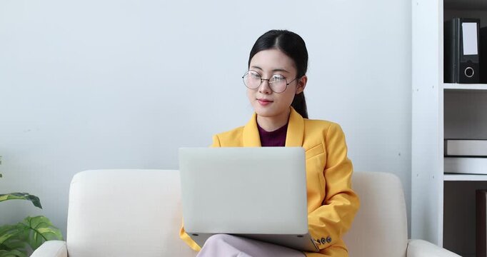 Young asian business woman focusing on her work, smiling at a laptop while sitting at a desk with documents and office supplies in a contemporary workspace