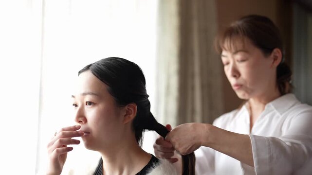 An adult carefully shapes a dark braid close to a seated woman head The slow motion highlights precise finger movements