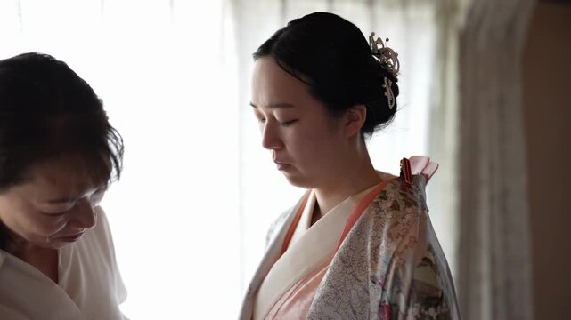 A dresser refines the collar of a light floral kimono on a young adult woman The close view emphasizes layered fabric and careful hands