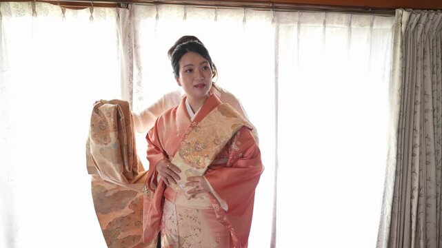 A dresser positions a decorative front panel over the obi area of an orange kimono The wearer maintains an upright formal pose