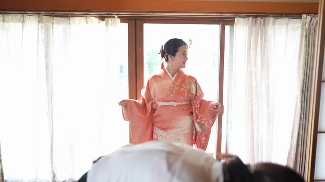 A woman gently extends the wide sleeves of her orange floral kimono while a dresser works at the waist Soft daylight illuminates the textile