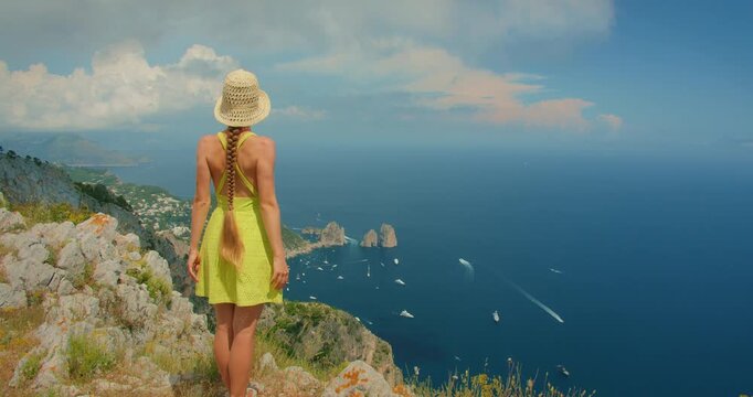 Woman standing on a cliff edge admiring the wide sea view with Faraglioni cliffs in Capri Italy