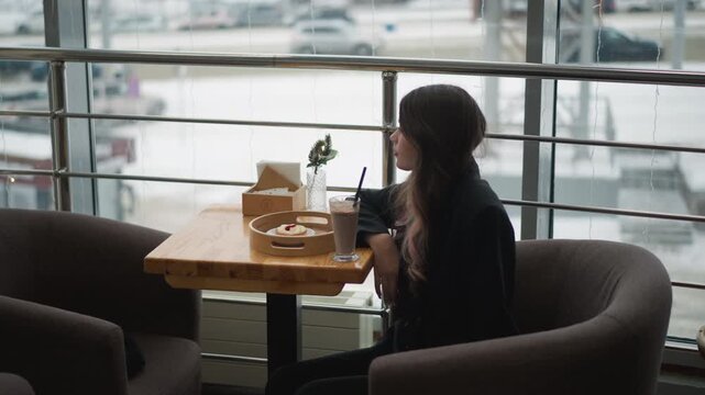 Young woman seated in airport lounge turns head toward camera while holding smartphone and takeaway cup, showing brief movement, calm expression, and reflective travel mood in soft daylight
