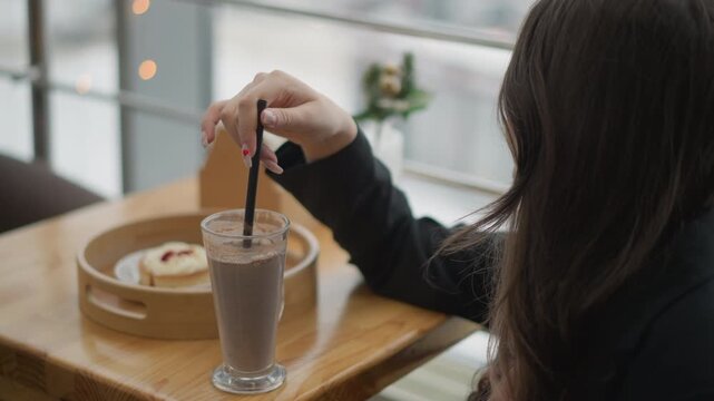 Close side view of young woman seated at wooden table in bright windowlit cafe, stirring iced coffee with metal straw; hand motion swirls foam, with condensation, soft daylight and relaxed mood