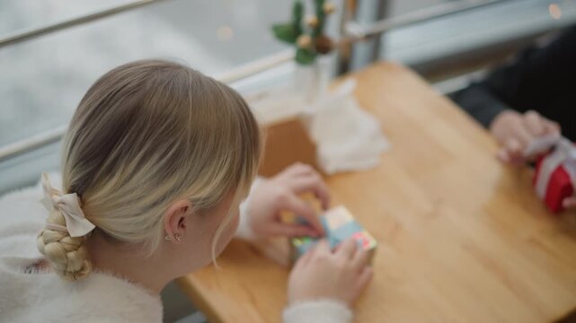 Rear above view shows young woman at wooden table actively turning colorful puzzle cube with both hands, focused expression, soft daylight, warm indoor scene, visible hand motion and quiet