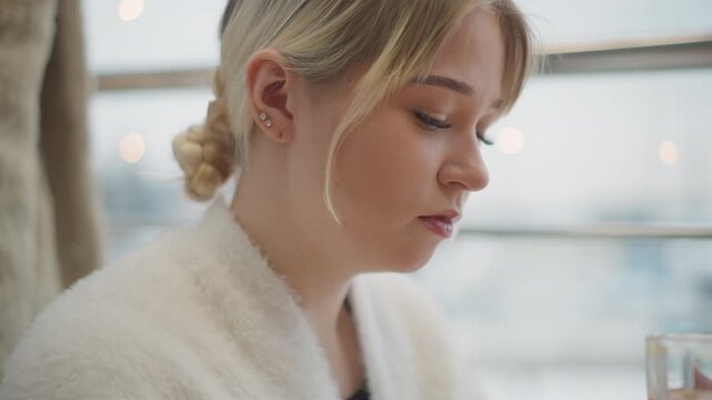 Indoor portrait shows young woman holding glass of orange beverage, taking gentle sip before quiet pause, head tilted down in soft daylight, creating calm reflective lifestyle scene