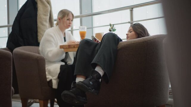Two adults sit facing each other in upholstered chairs inside modern cafe lounge, one leaning forward with drink while other reclines with legs raised, showing relaxed conversation, comfort, and