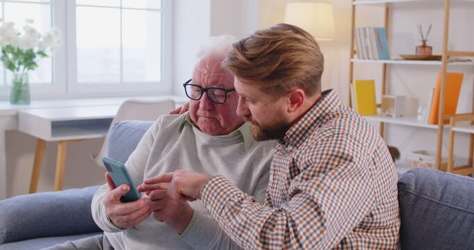 Adult son explains and teaches his elderly father how to use mobile app on smartphone. Millennial son with older father sitting on sofa where son is friendly explaining functions of modern technology.
