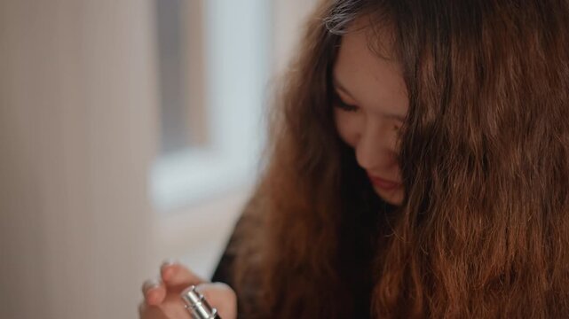 Long curly-haired young woman tilts head and dispenses liquid from small dropper bottle onto fingertip, showing careful hand movement, focused attention, soft indoor light, and natural beauty routine
